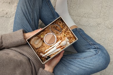 Photo of Woman holding Christmas gift box with makeup brushes and cosmetics at home, above view