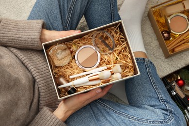 Photo of Woman holding Christmas gift box with makeup brushes and cosmetics at home, above view