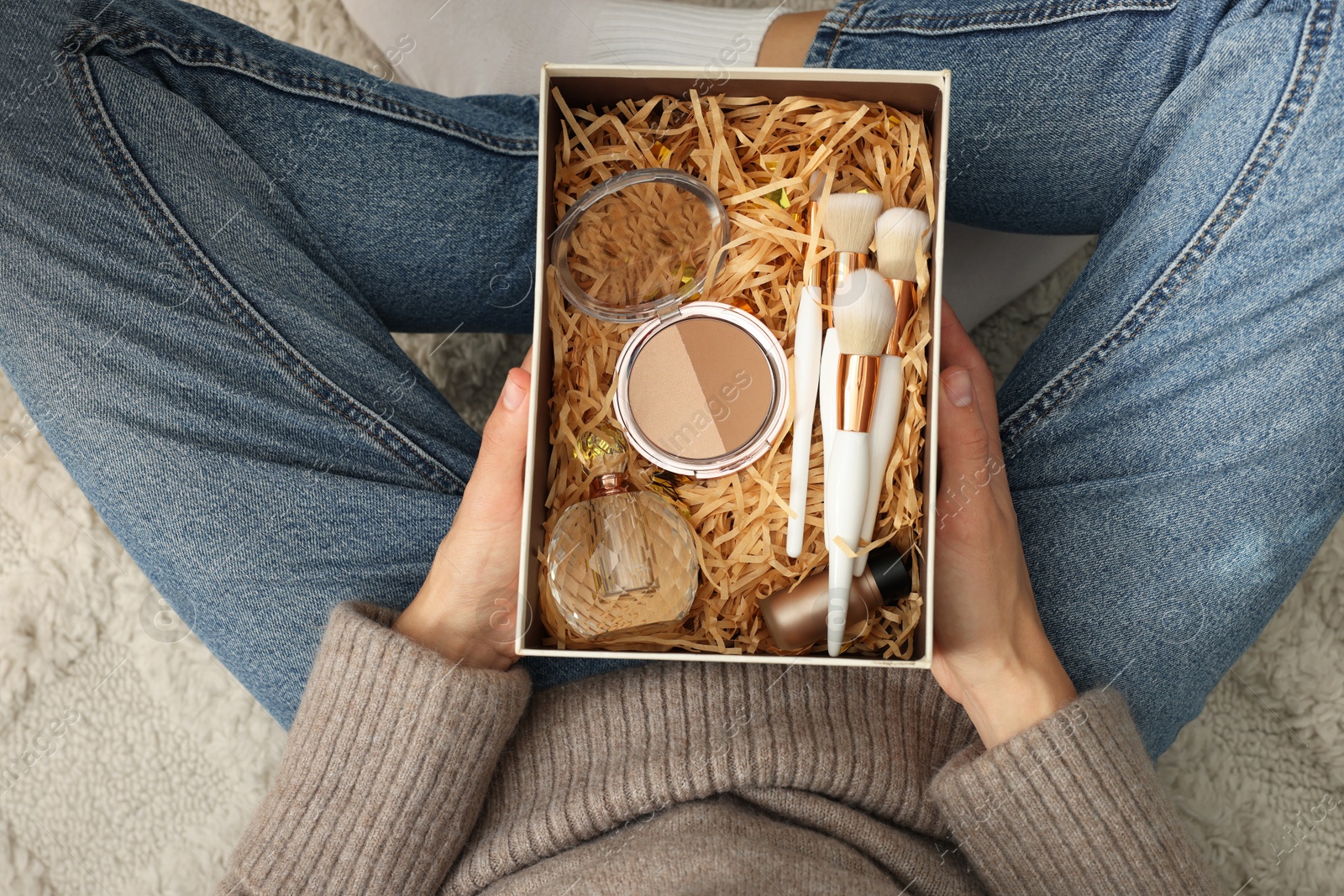 Photo of Woman holding Christmas gift box with makeup brushes and cosmetics at home, above view