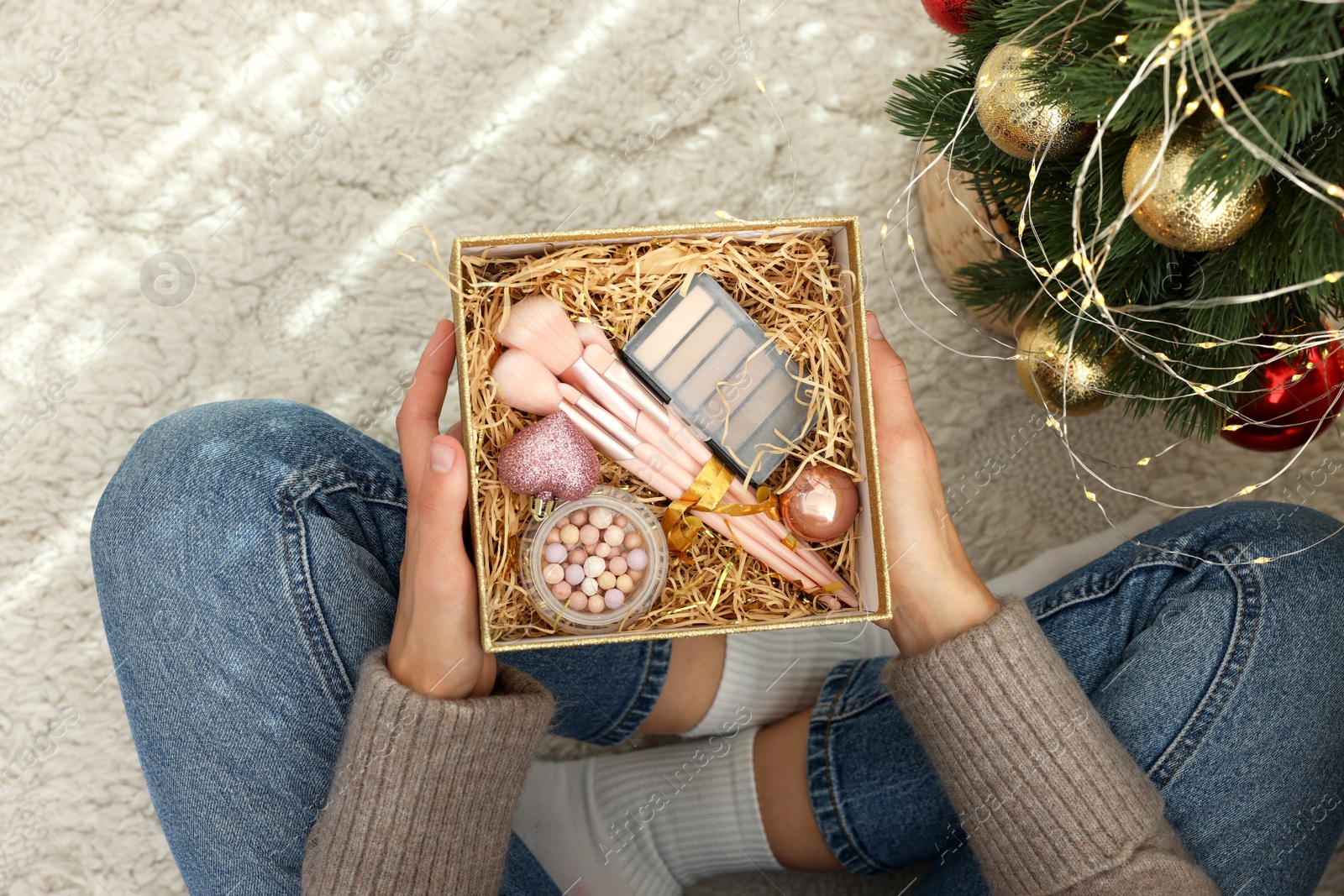 Photo of Woman holding Christmas gift box with makeup brushes and cosmetics at home, above view