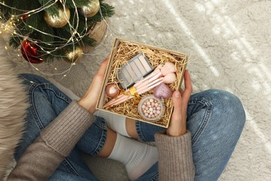 Photo of Woman holding Christmas gift box with makeup brushes and cosmetics at home, above view