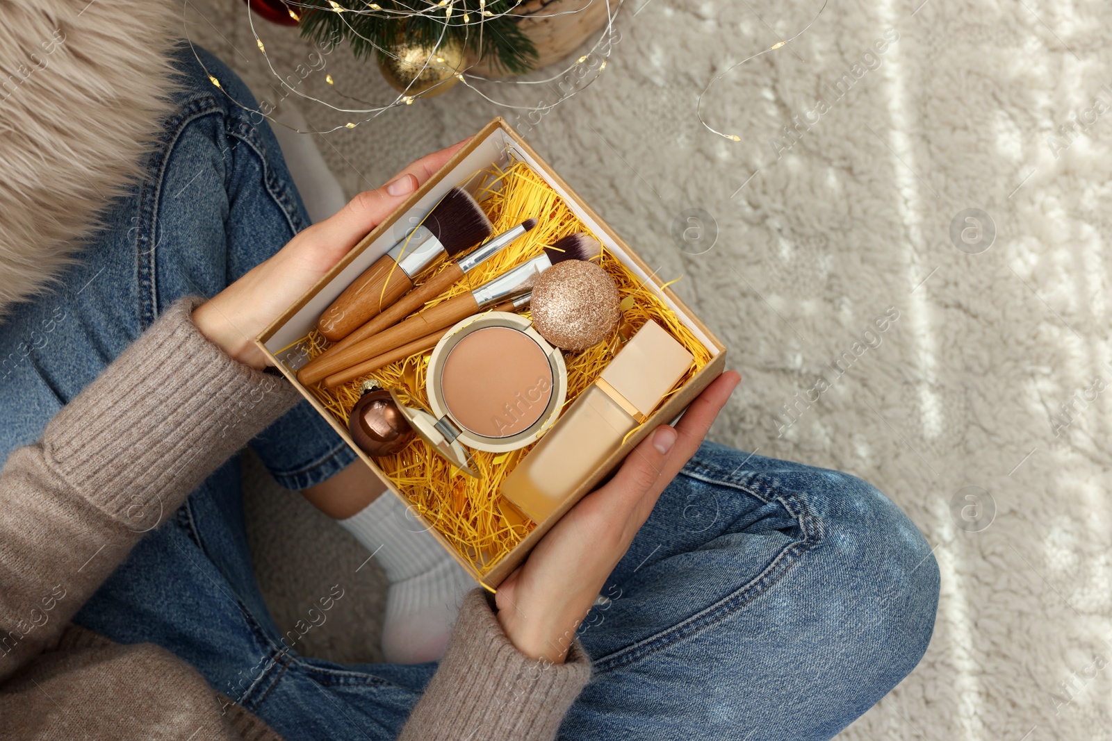 Photo of Woman holding Christmas gift box with makeup brushes and cosmetics at home, above view