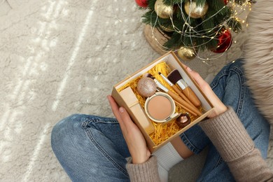 Photo of Woman holding Christmas gift box with makeup brushes and cosmetics at home, above view