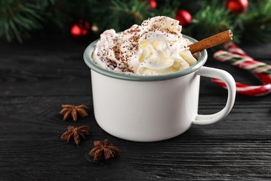 Photo of Tasty hot cocoa drink with whipped cream in mug and spices on black wooden table, closeup