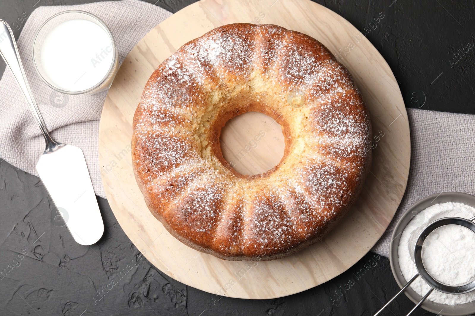 Photo of Freshly baked sponge cake with powdered sugar, milk and server on black table, top view