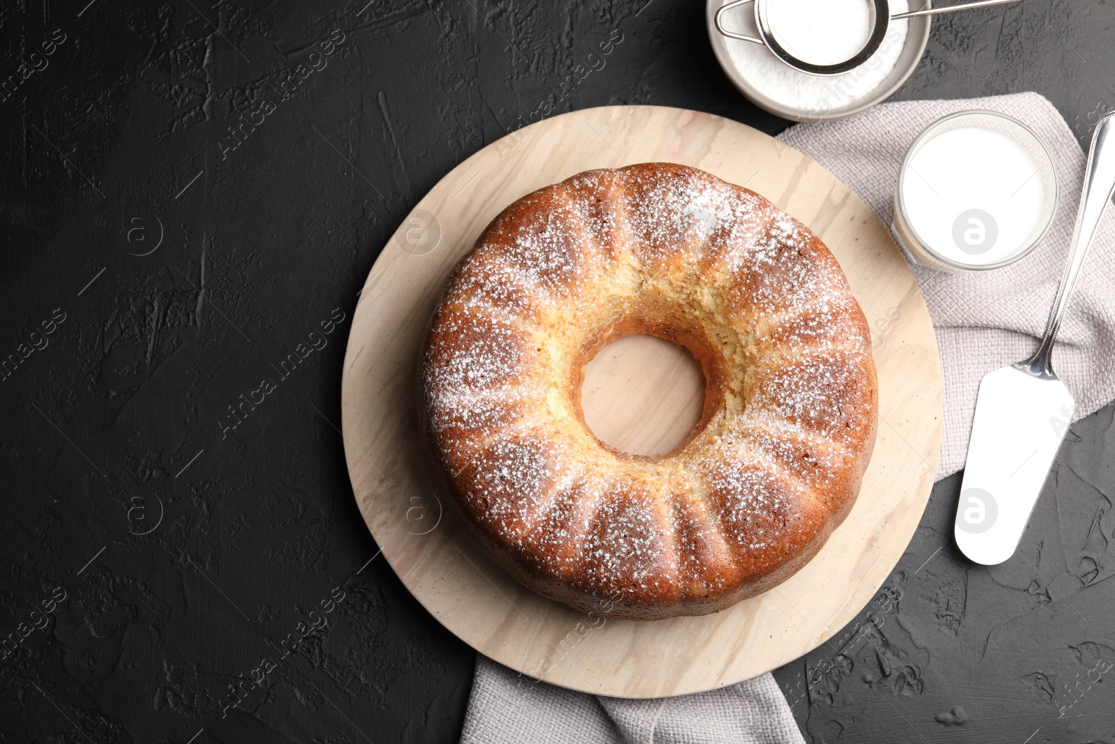 Photo of Freshly baked sponge cake with powdered sugar, milk and server on black table, top view