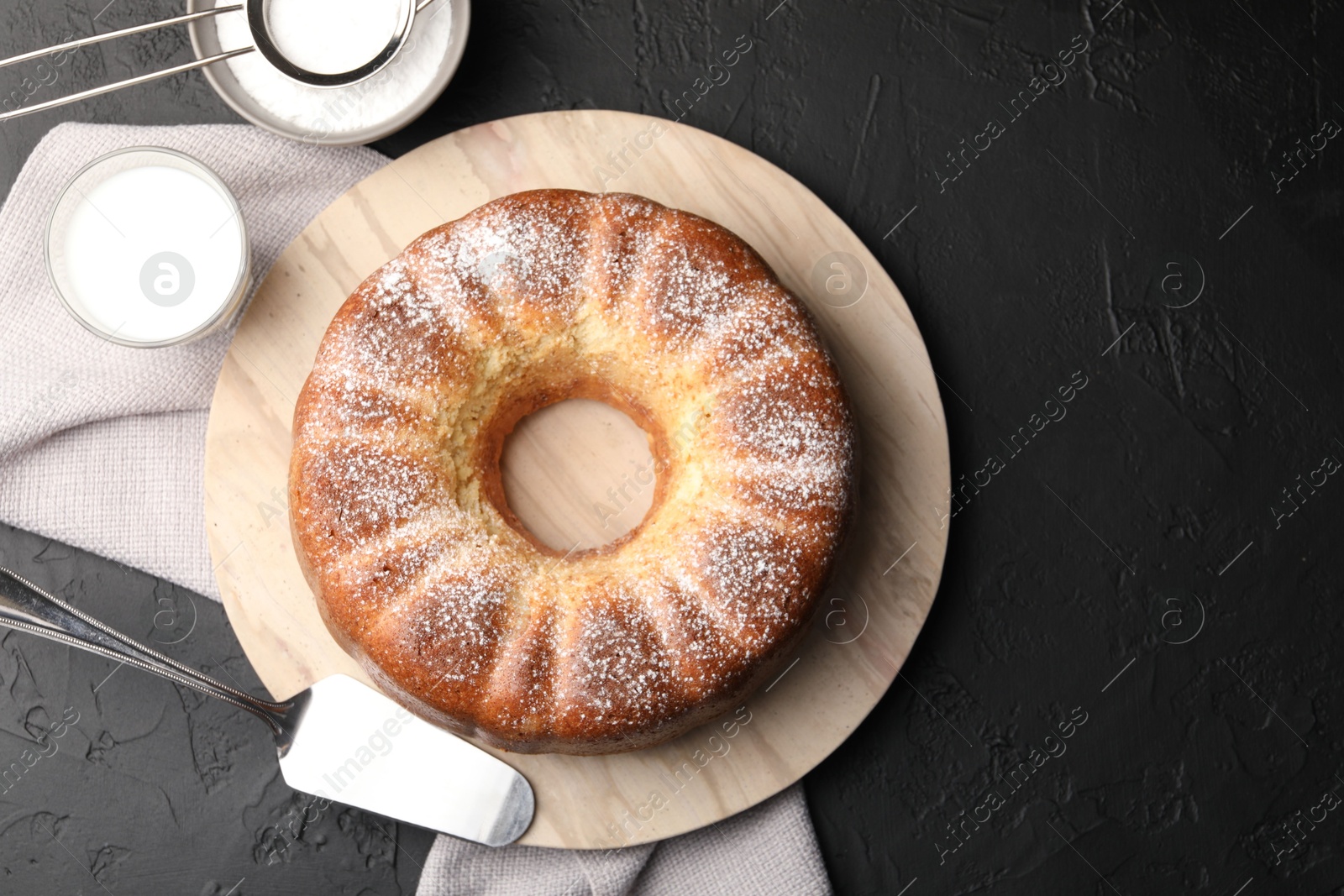 Photo of Freshly baked sponge cake with powdered sugar, milk and server on black table, top view