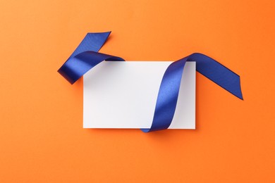 Blank card and blue ribbon on orange background, top view