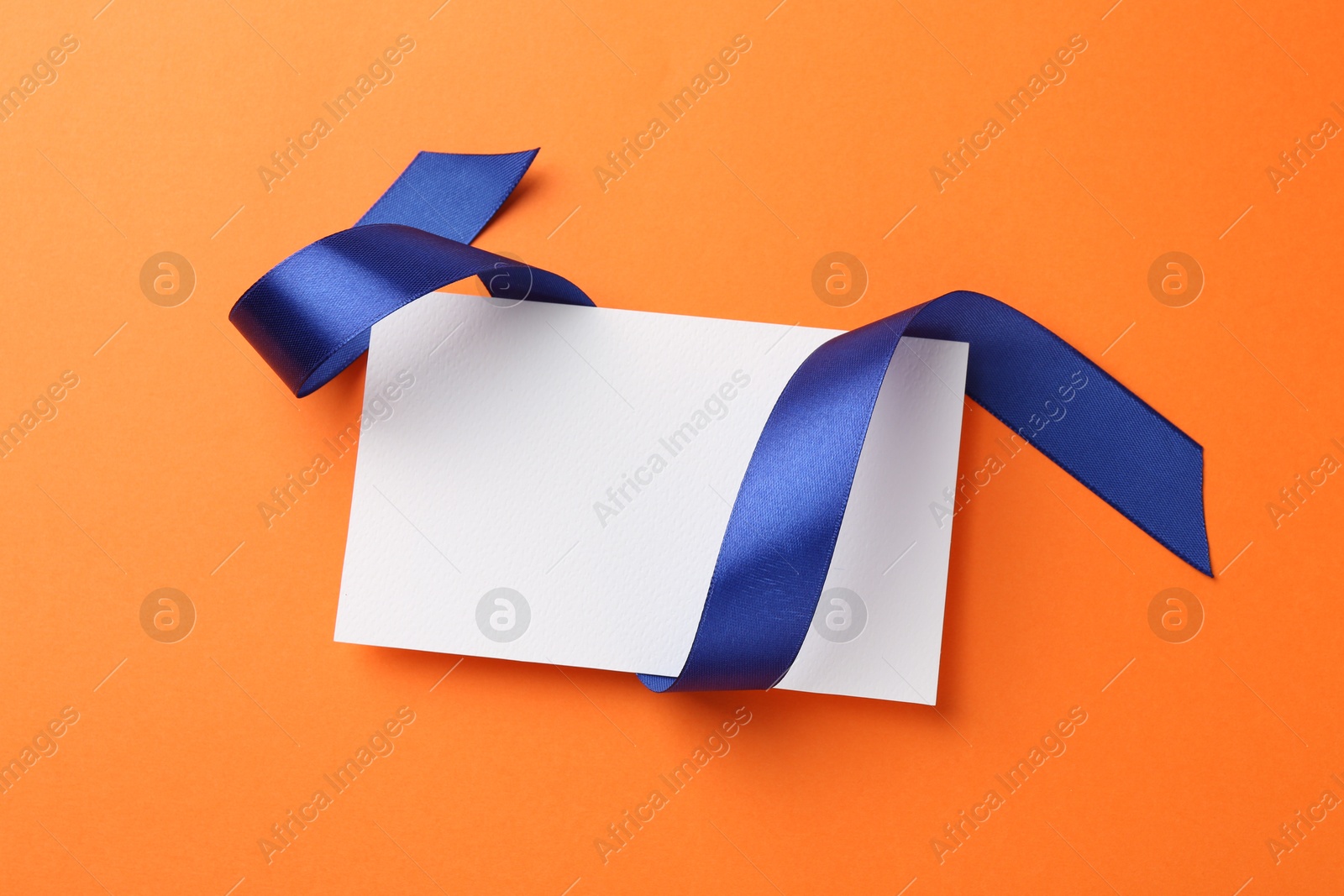 Photo of Blank card and blue ribbon on orange background, top view