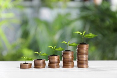 Image of Stacked coins with green seedlings on white table against blurred background, space for text