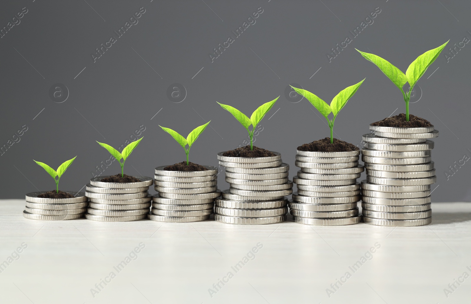 Image of Stacked coins with green seedlings on white table against grey background