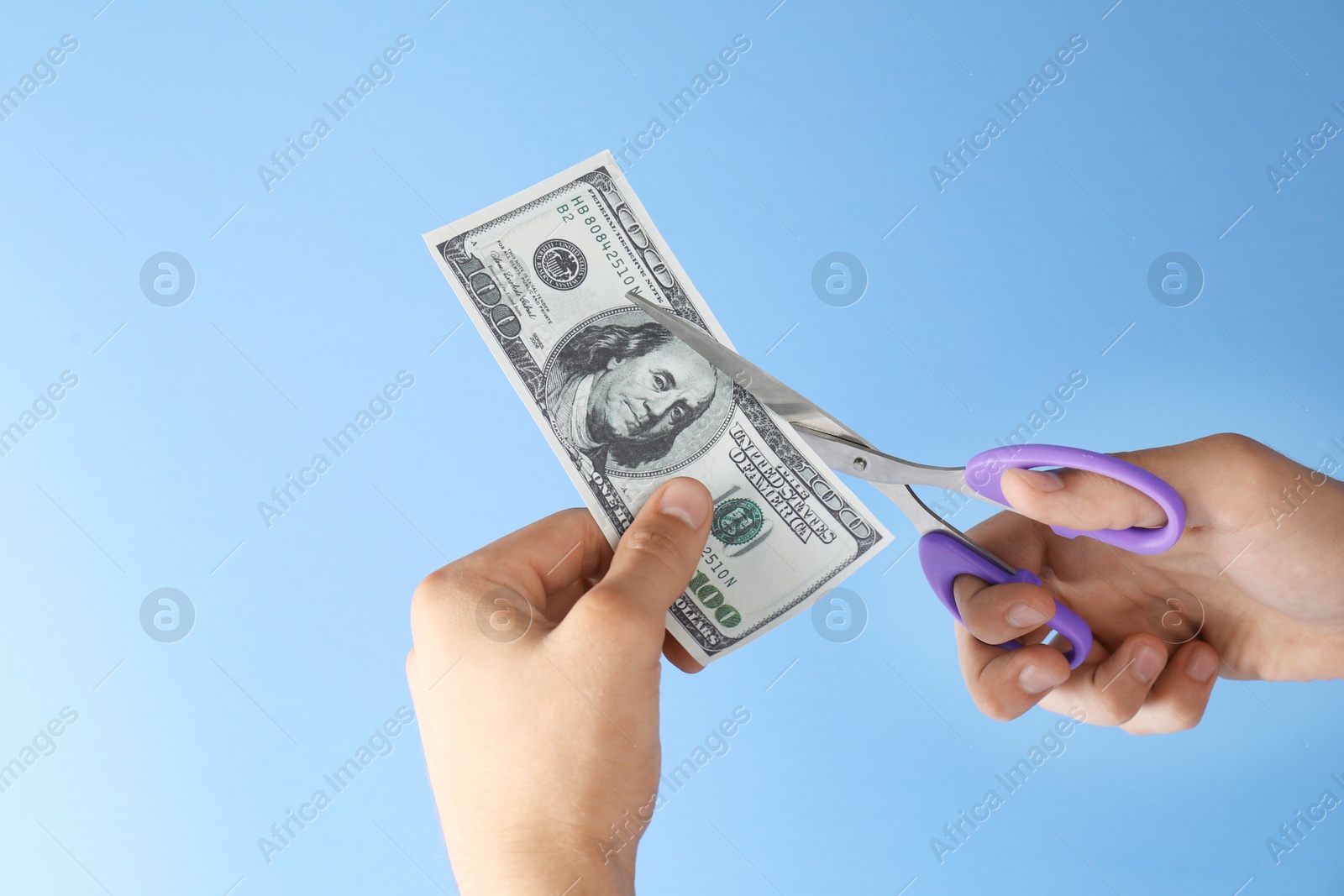 Photo of Budgeting. Man cutting dollar banknote with scissors on light blue background, closeup