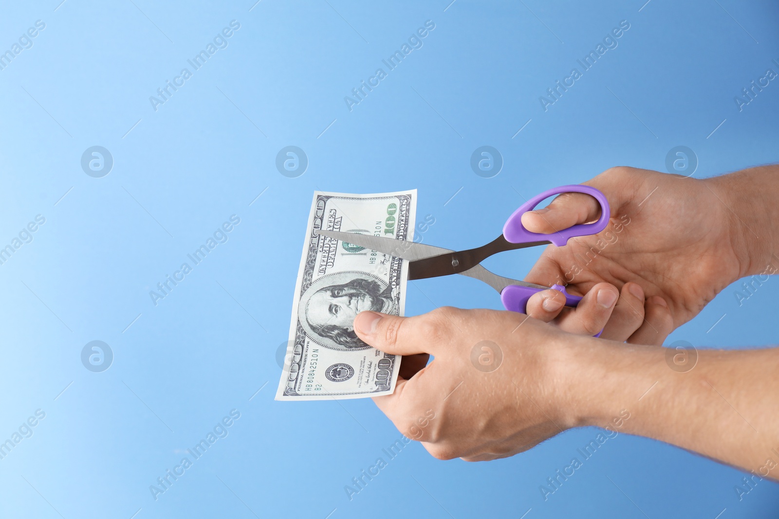 Photo of Budgeting. Man cutting dollar banknote with scissors on light blue background, closeup. Space for text