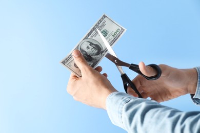 Photo of Budgeting. Man cutting dollar banknote with scissors on light blue background, closeup. Space for text