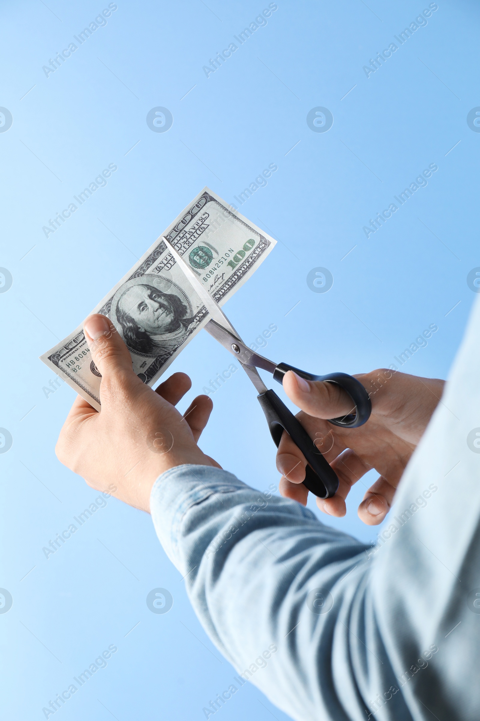 Photo of Budgeting. Man cutting dollar banknote with scissors on light blue background, closeup