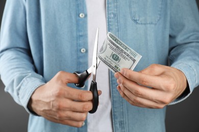 Photo of Budgeting. Man cutting dollar banknote with scissors on dark grey background, closeup