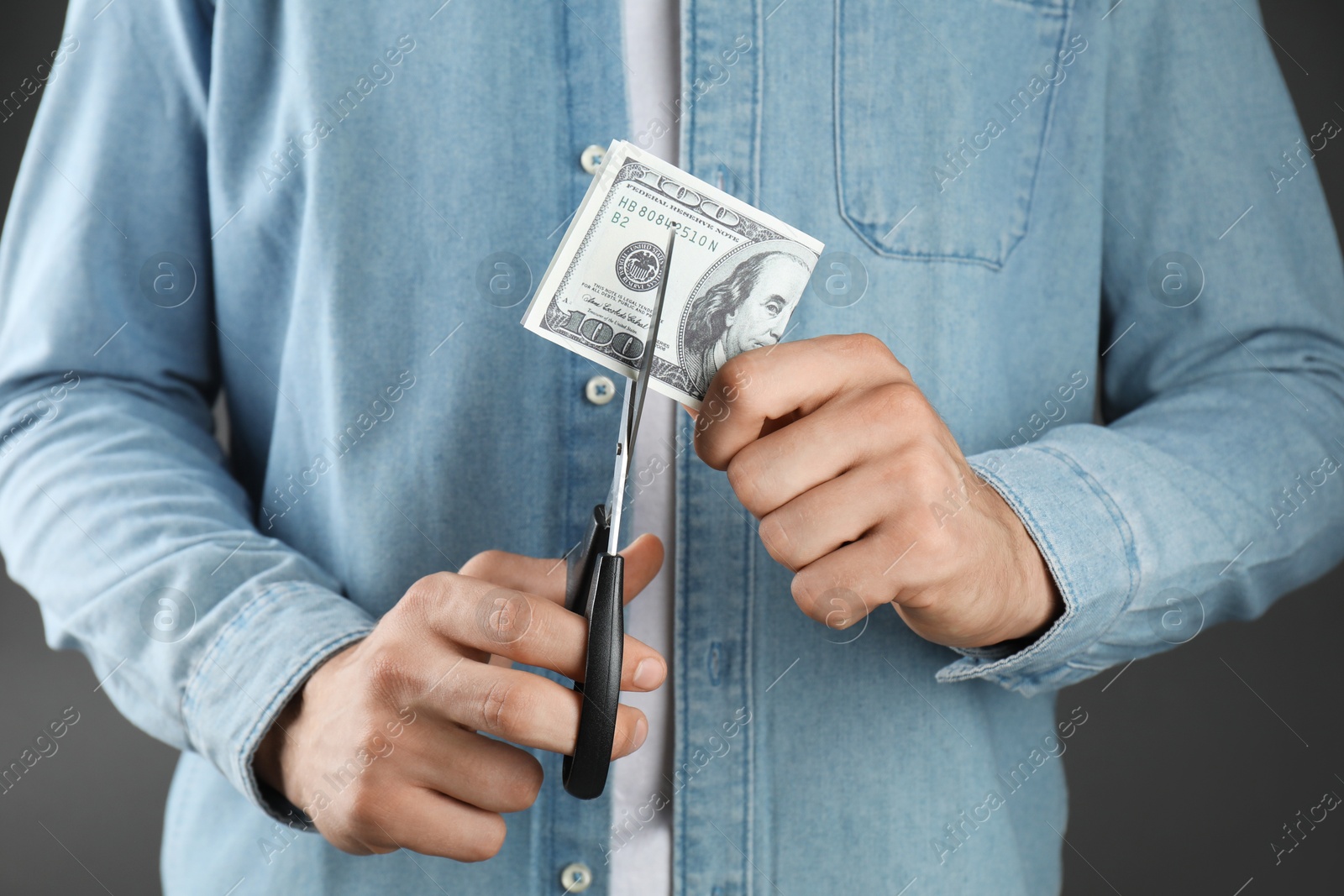 Photo of Budgeting. Man cutting dollar banknote with scissors on dark grey background, closeup