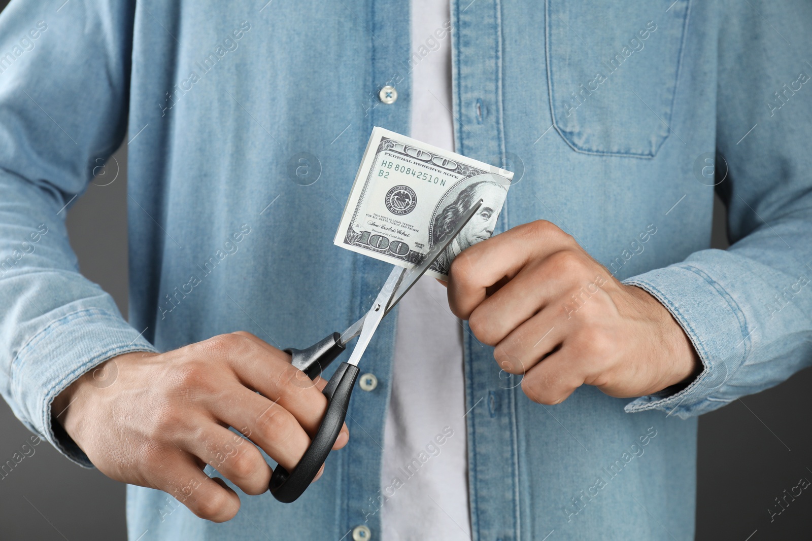 Photo of Budgeting. Man cutting dollar banknote with scissors on dark grey background, closeup