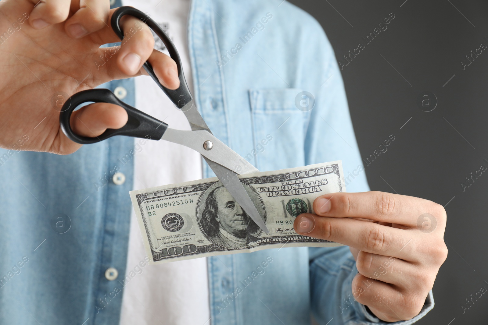 Photo of Budgeting. Man cutting dollar banknote with scissors on dark grey background, closeup