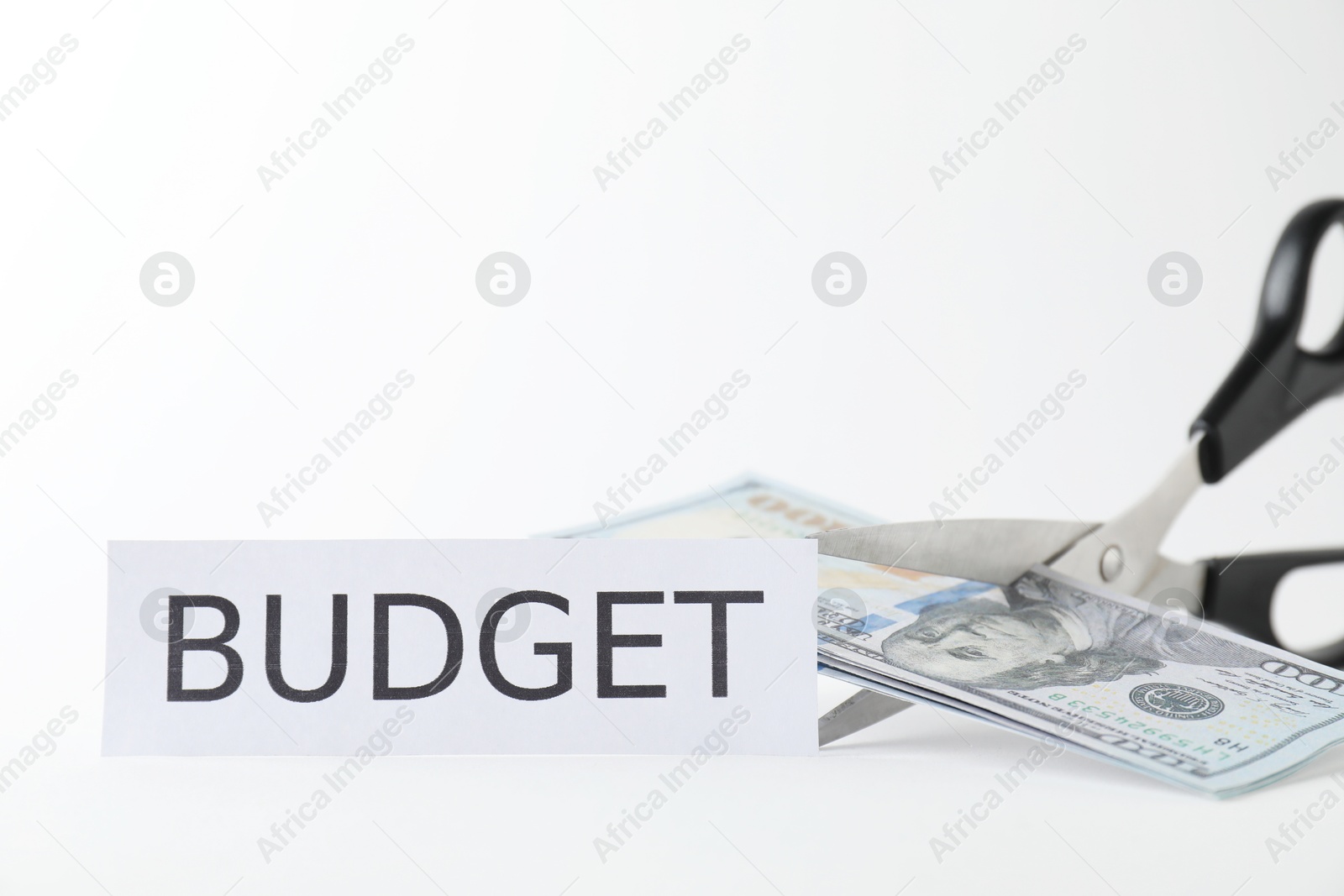 Photo of Cutting dollar banknotes with scissors and paper with word Budget on white background, closeup. Space for text