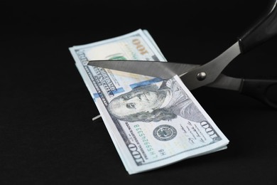 Photo of Budgeting. Cutting dollar banknotes with scissors on black background, closeup