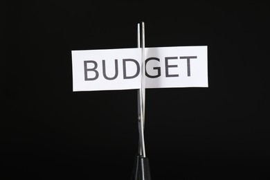 Photo of Cutting paper with word Budget with scissors on black background, closeup