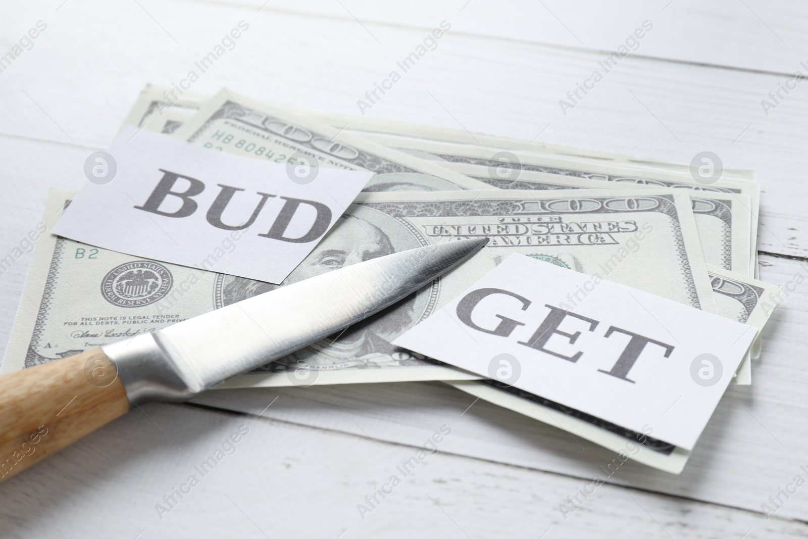 Photo of Cut paper with word Budget, dollar banknotes and knife on white wooden table, closeup