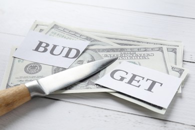 Photo of Cut paper with word Budget, dollar banknotes and knife on white wooden table, closeup