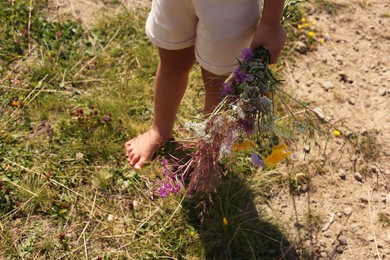 Photo of Little girl with bouquet of wildflowers walking barefoot outdoors on sunny day, closeup. Child enjoying beautiful nature