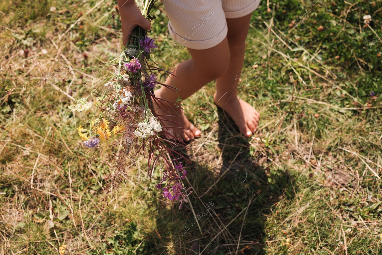 Photo of Little girl with bouquet of wildflowers walking barefoot outdoors on sunny day, closeup. Child enjoying beautiful nature