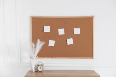 Cork board with blank paper notes, alarm clock and decorative branches in vase on wooden table