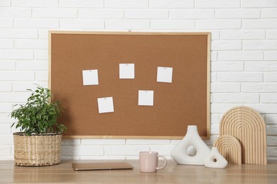 Photo of Cork board with blank paper notes, laptop and decorative elements on wooden table near white brick wall