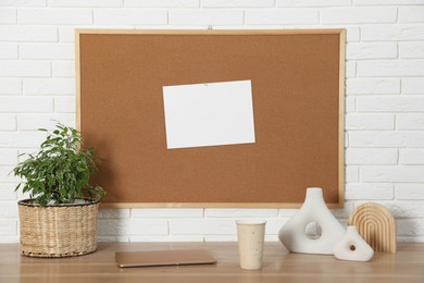 Photo of Cork board with blank paper note, laptop and decorative elements on wooden table near white brick wall