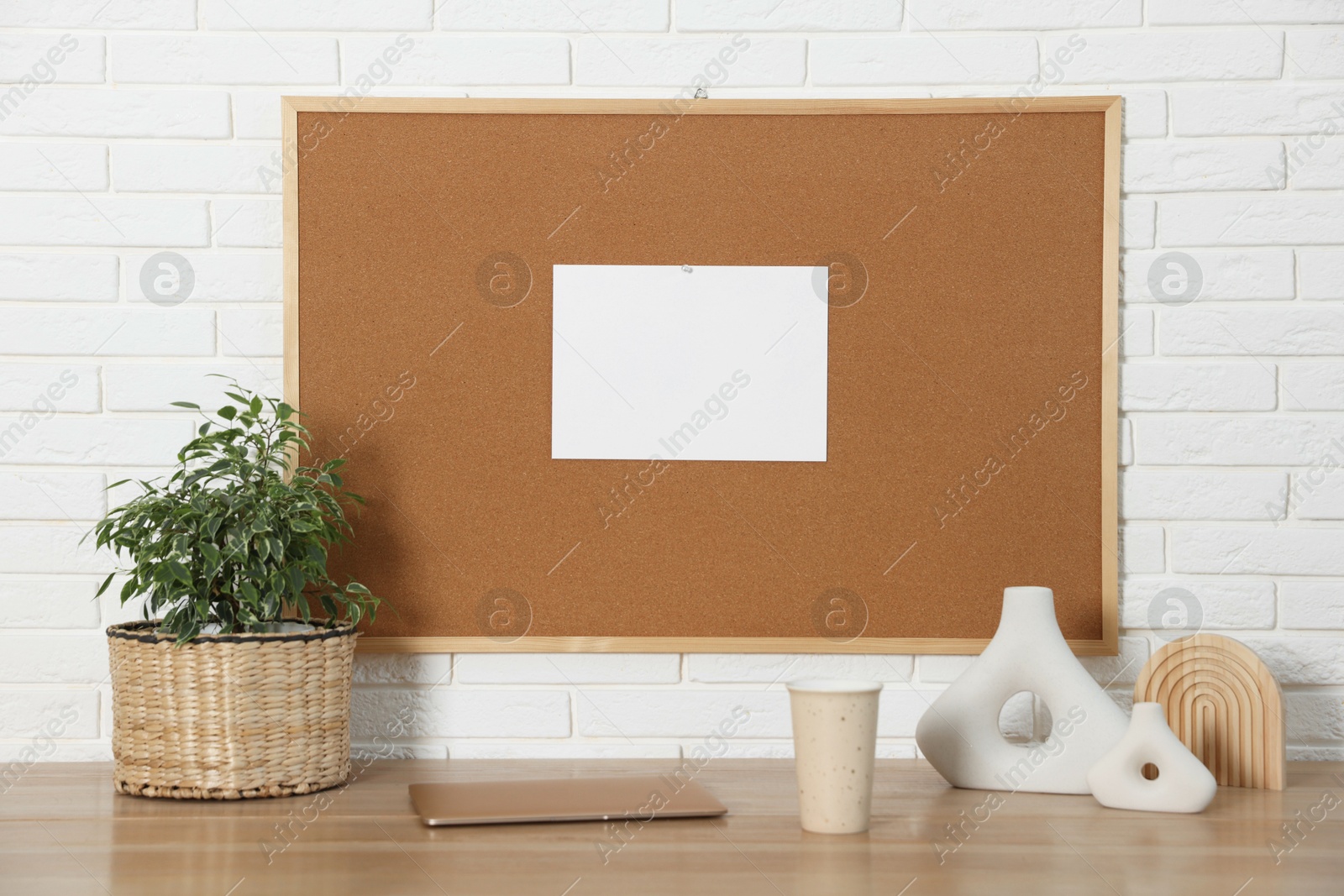 Photo of Cork board with blank paper note, laptop and decorative elements on wooden table near white brick wall