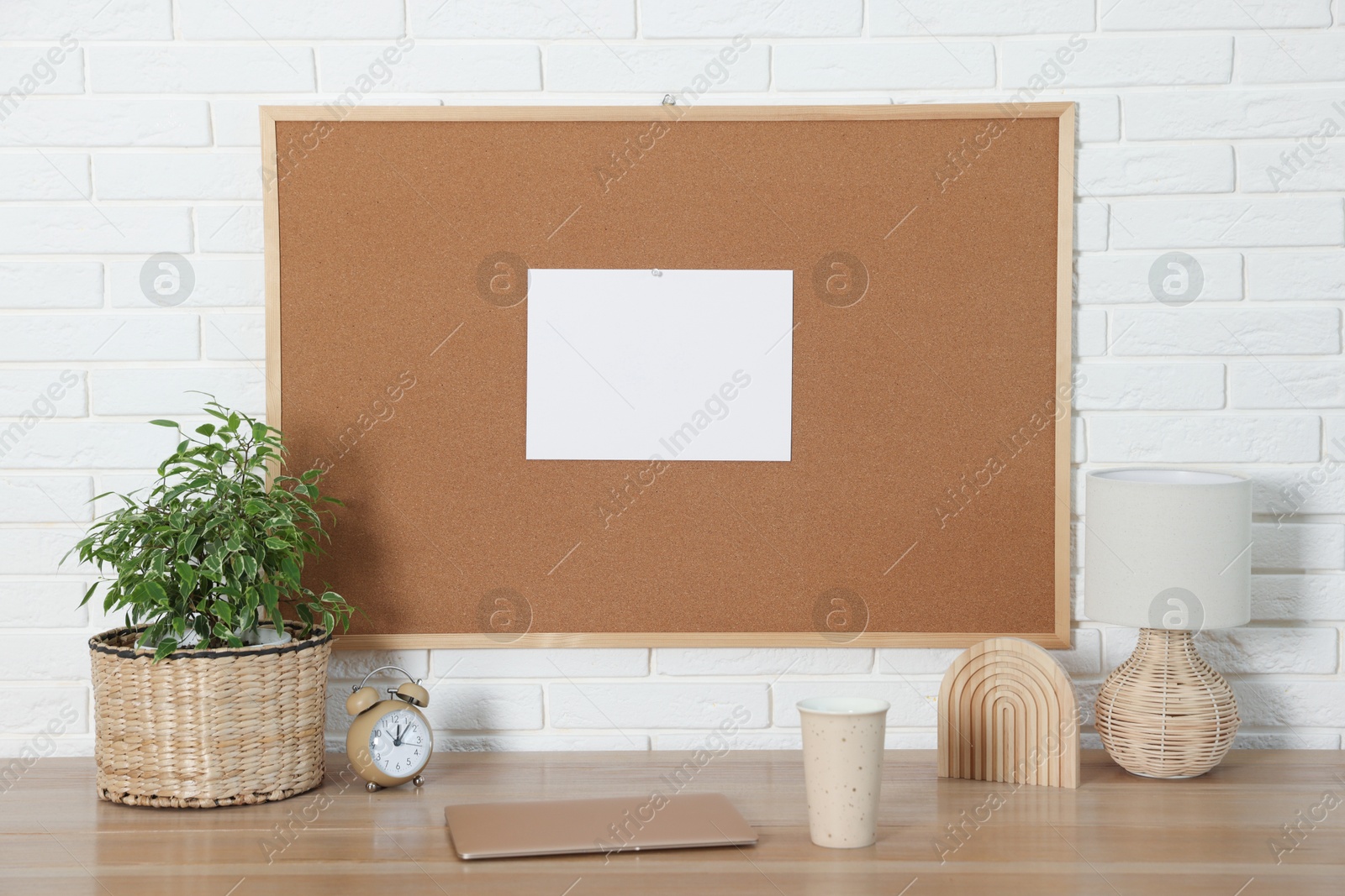 Photo of Cork board with blank paper note, laptop and decorative elements on wooden table near white brick wall