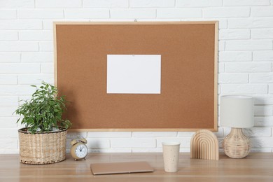 Cork board with blank paper note, laptop and decorative elements on wooden table near white brick wall