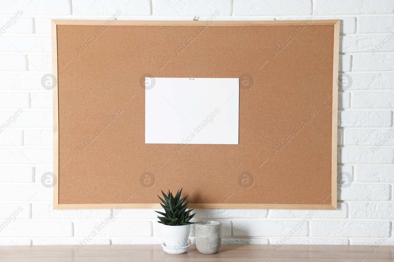 Photo of Cork board with blank paper note, houseplant and scented candle on wooden table near white brick wall