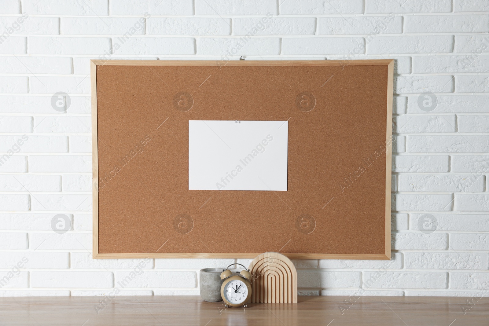 Photo of Cork board with blank paper note and decorative elements on wooden table near white brick wall