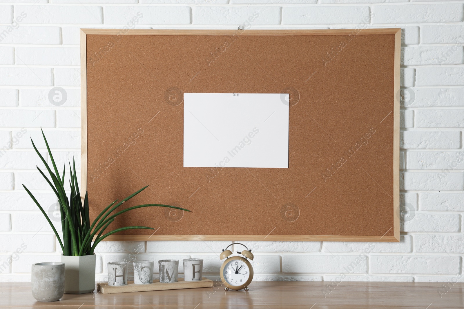 Photo of Cork board with blank paper note and decorative elements on wooden table near white brick wall