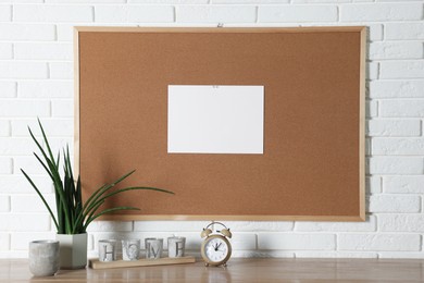 Photo of Cork board with blank paper note and decorative elements on wooden table near white brick wall