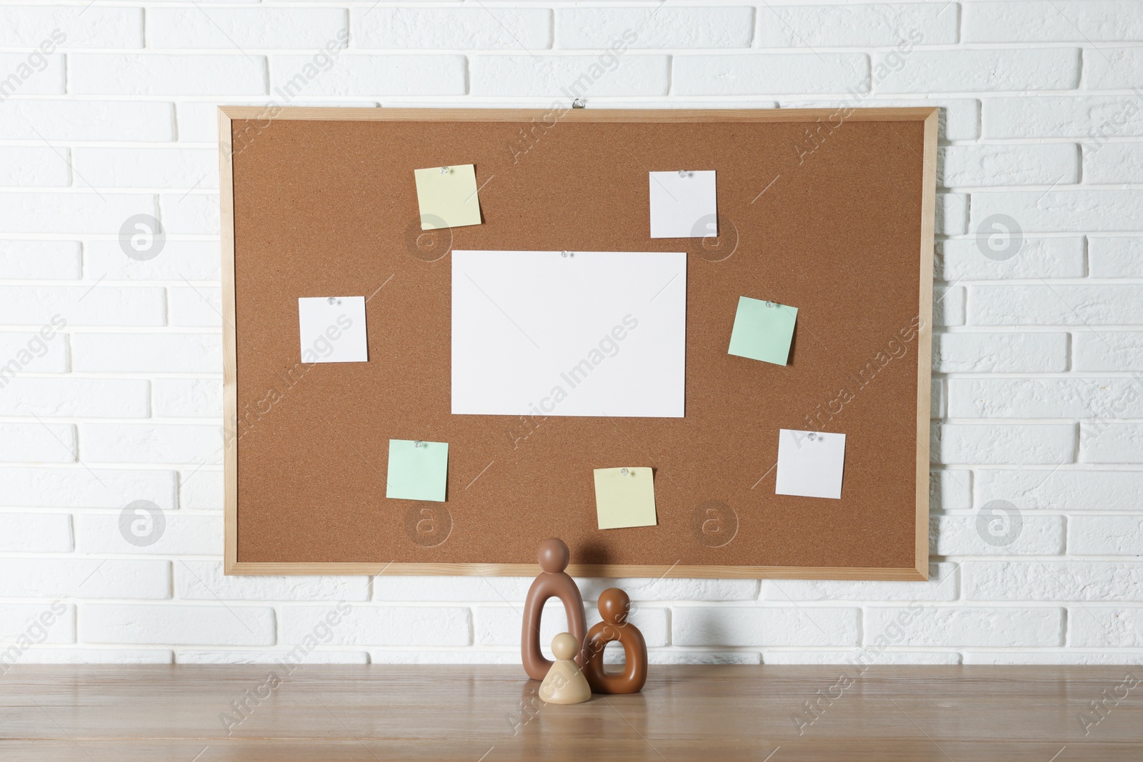 Photo of Cork board with blank paper notes and decorative figures on wooden table near white brick wall