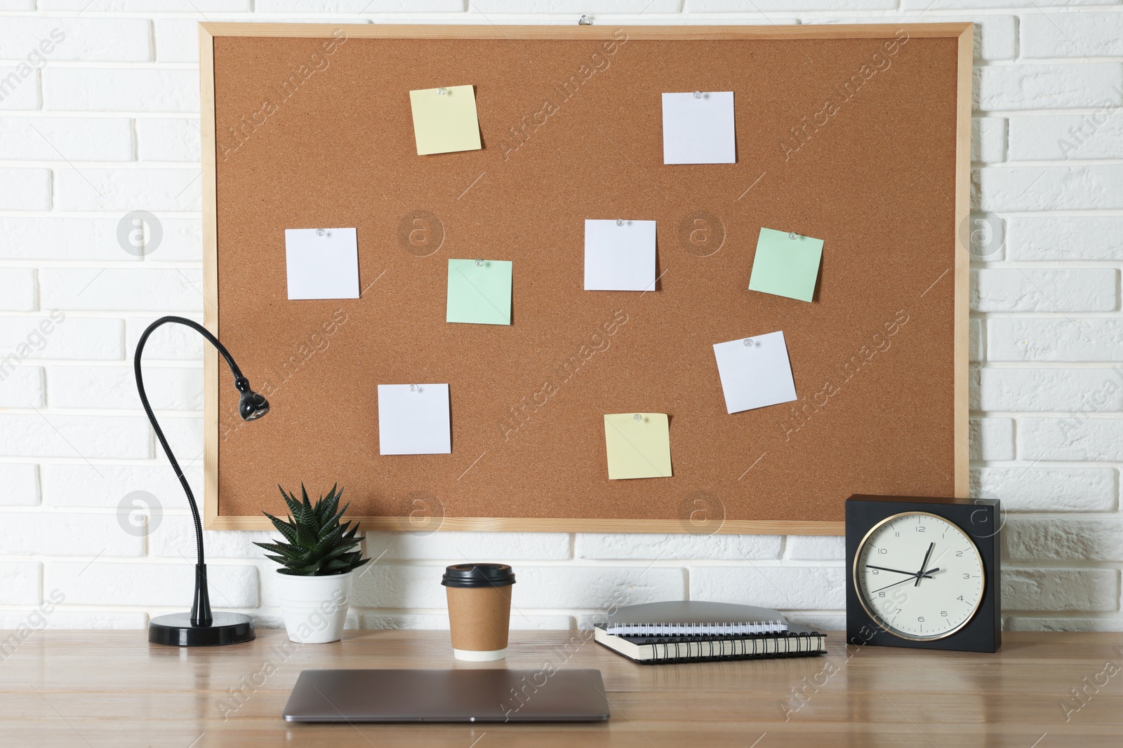 Photo of Cork board with blank paper notes, laptop, lamp, alarm clock and notebooks on wooden table near white brick wall