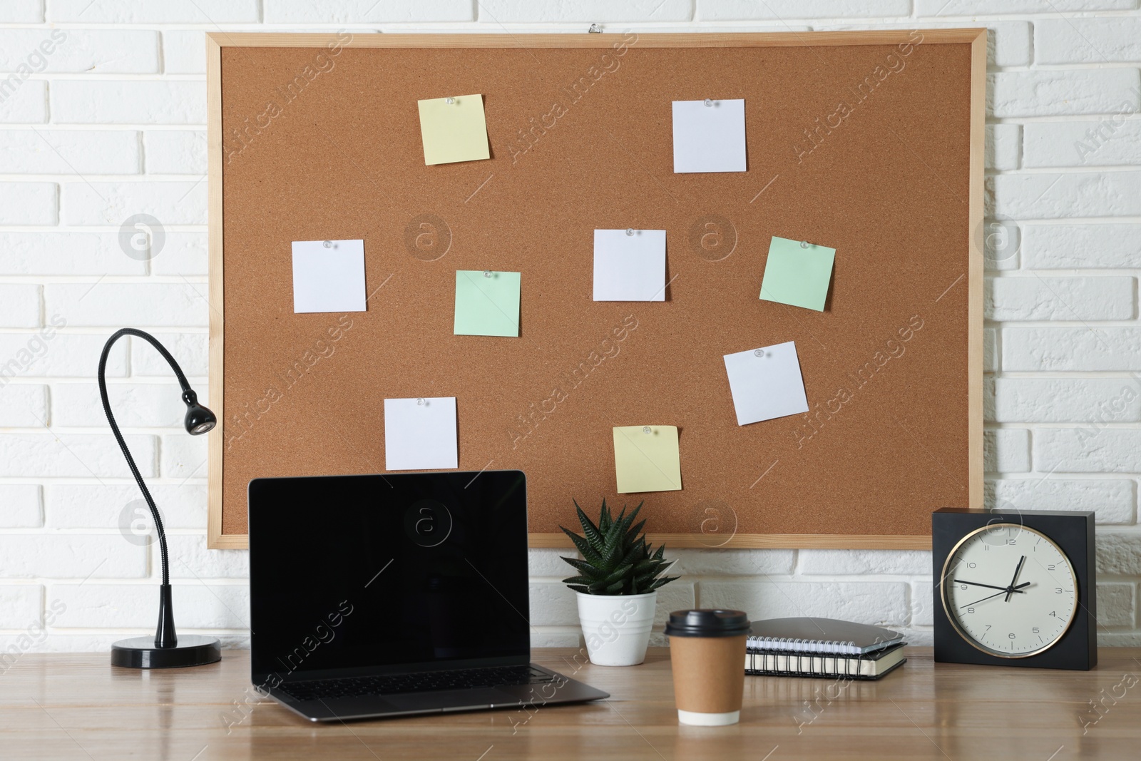 Photo of Cork board with blank paper notes, laptop, lamp, alarm clock and notebooks on wooden table near white brick wall