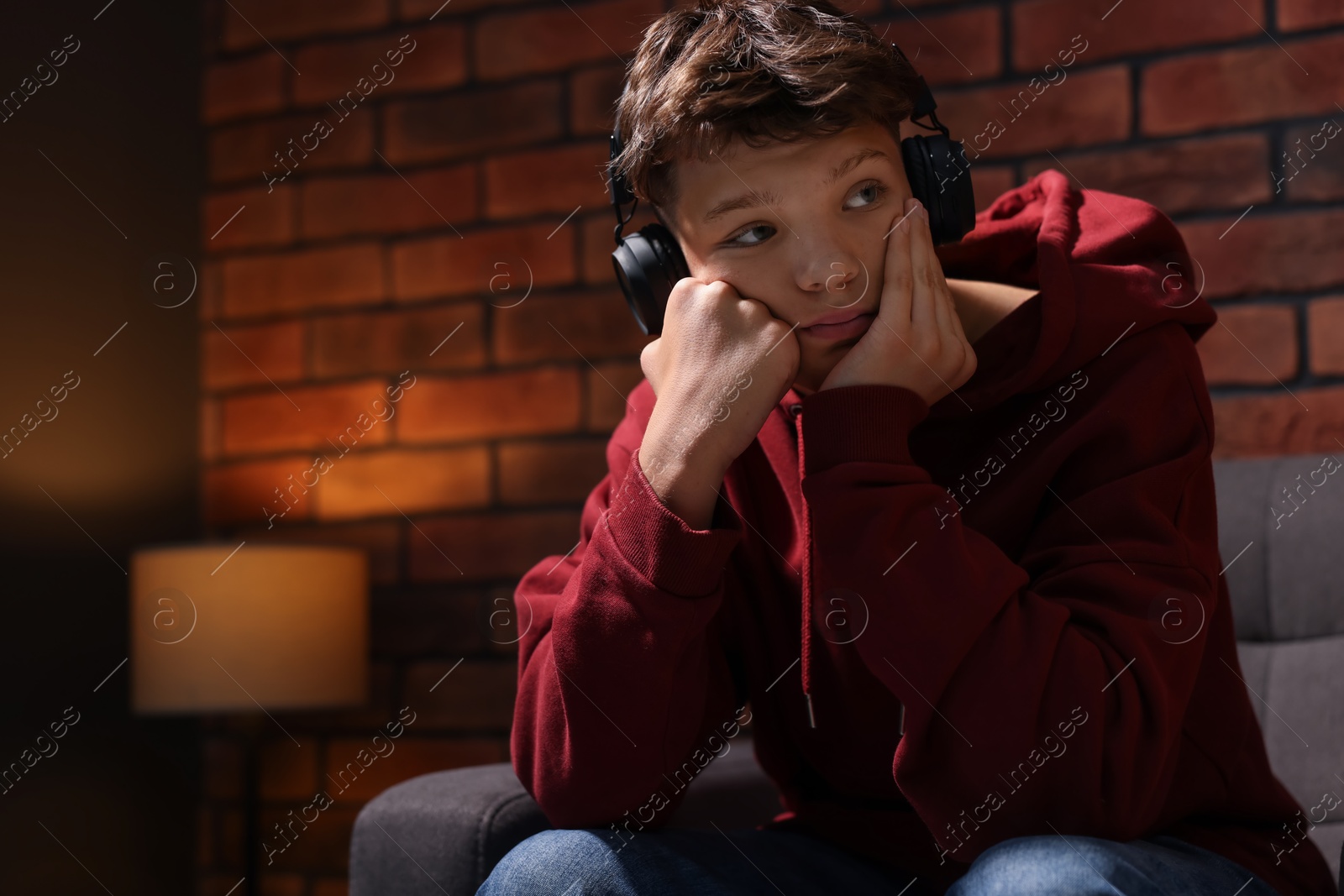 Photo of Loneliness concept. Sad teenage boy in headphones listening to music on armchair near brick wall