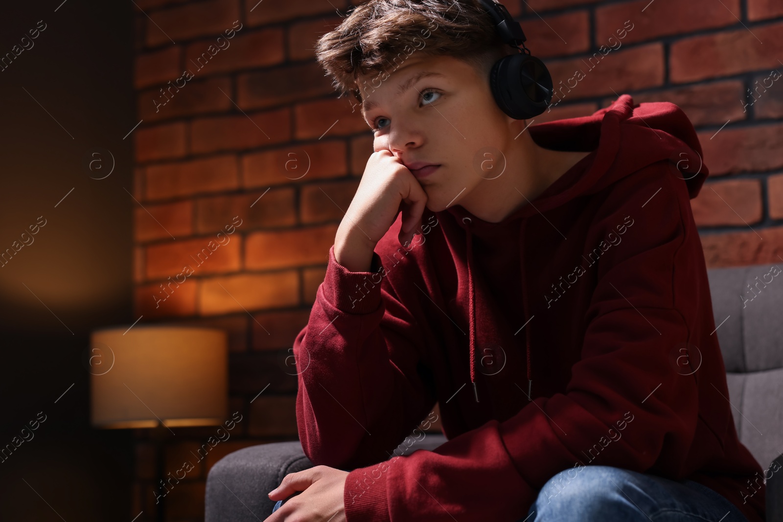 Photo of Loneliness concept. Sad teenage boy in headphones listening to music on armchair near brick wall