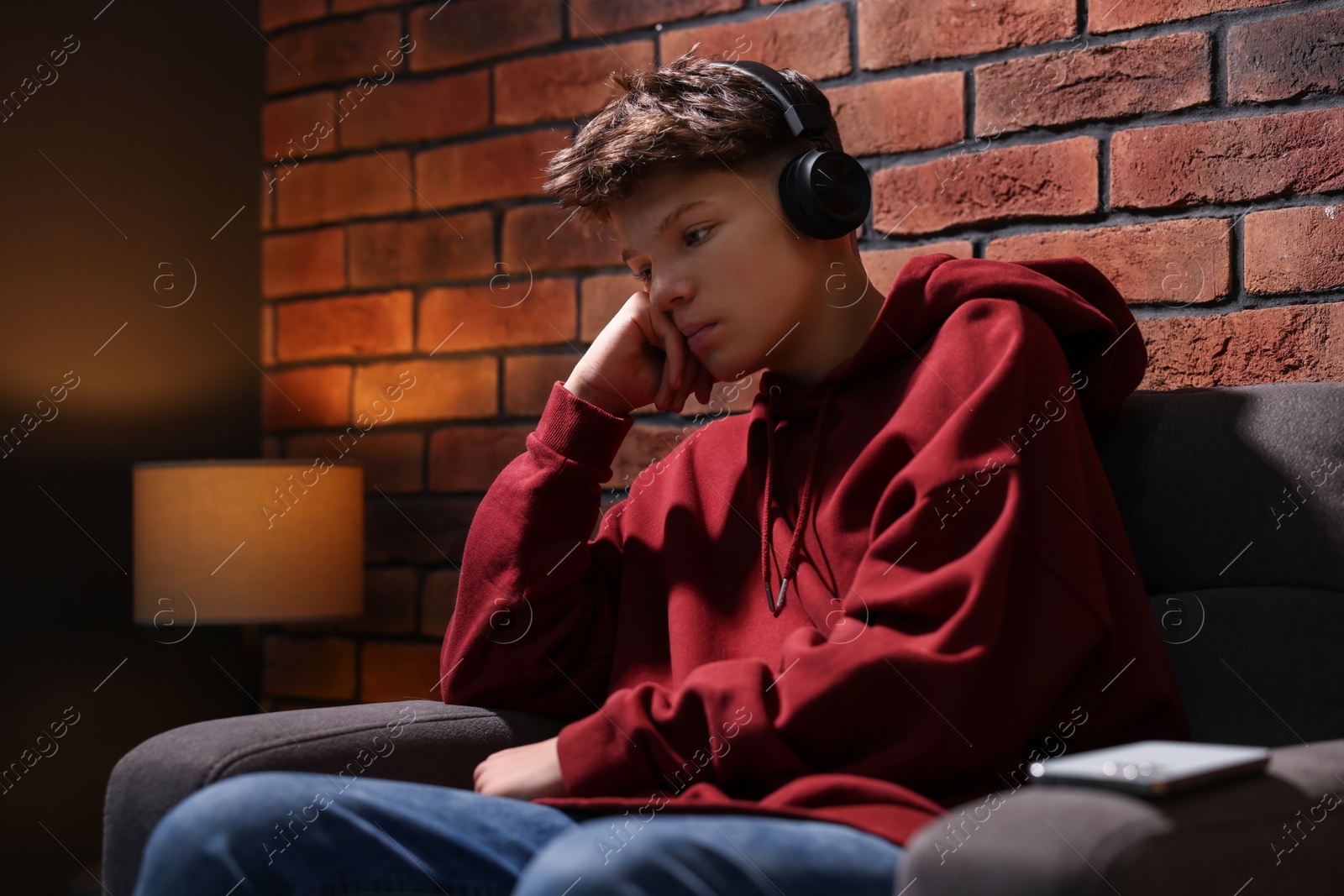 Photo of Loneliness concept. Sad teenage boy in headphones listening to music on armchair near brick wall