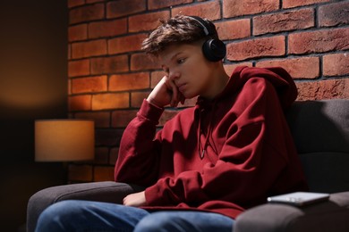 Photo of Loneliness concept. Sad teenage boy in headphones listening to music on armchair near brick wall