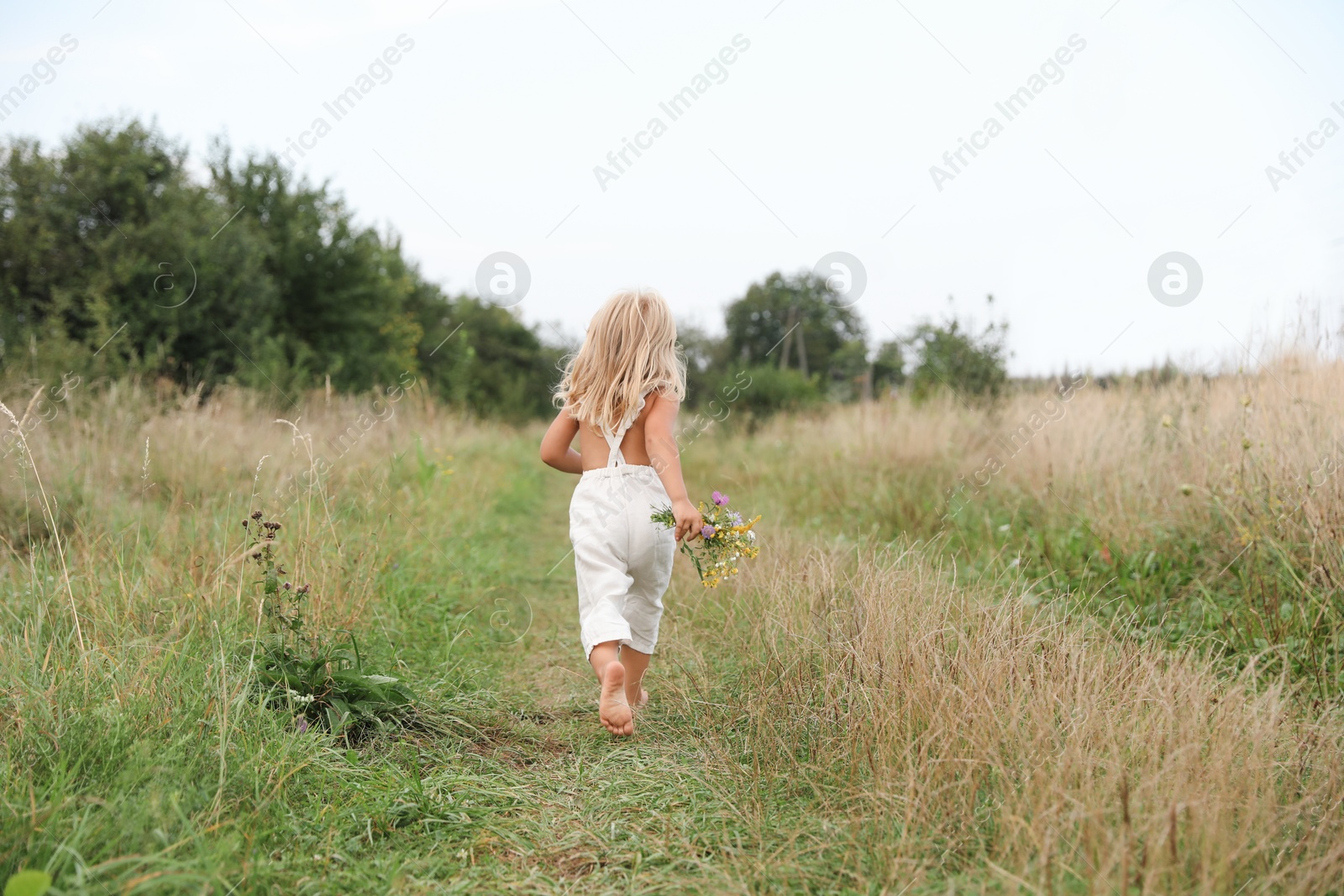 Photo of Little girl with flowers walking barefoot at meadow, back view. Child enjoying beautiful nature