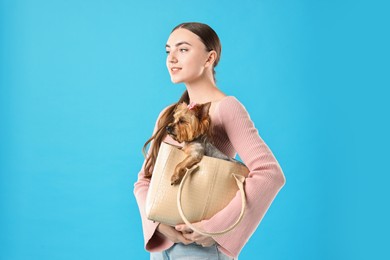 Beautiful young woman holding bag with cute Yorkshire Terrier dog on light blue background