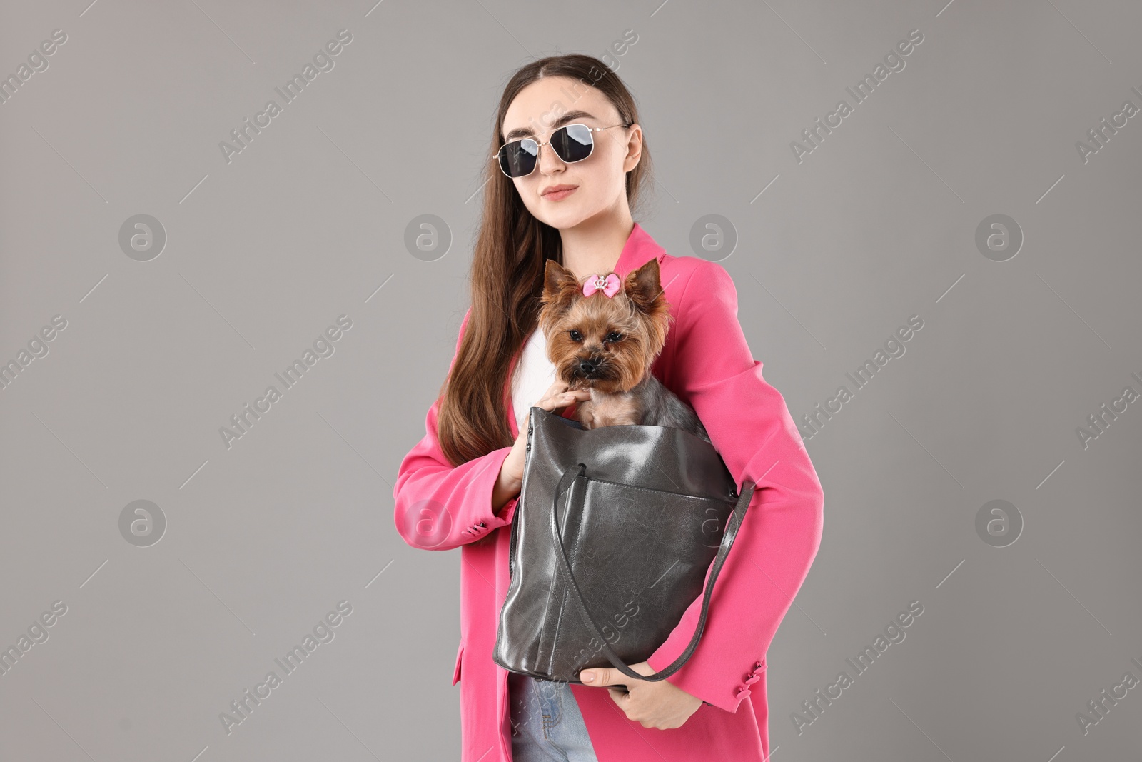 Photo of Woman in sunglasses carrying cute Yorkshire Terrier dog in bag on grey background